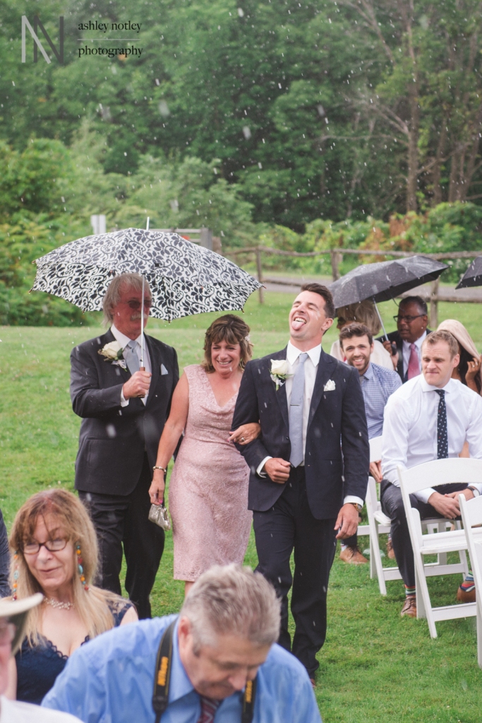 Groom walking down the aisle with his parents with his tongue sticking out while raining